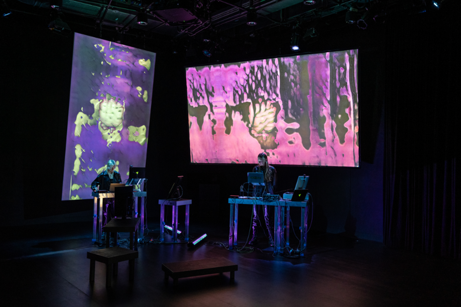 Photography of a stage situation. Allapopp and Janne Kummer are standing at desks with computers on them. In the background there are two screens on which the live mappings are projected.