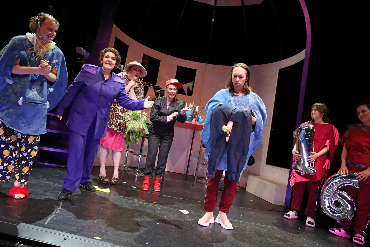 Several people are standing on a stage, their attention is directed towards a person standing somewhat in the foreground, who is holding a pair of trousers in his hand from which a sock is sticking out, reminiscent of an erect penis. Two people on the far right of the picture are holding a silver one and a six as a balloon.