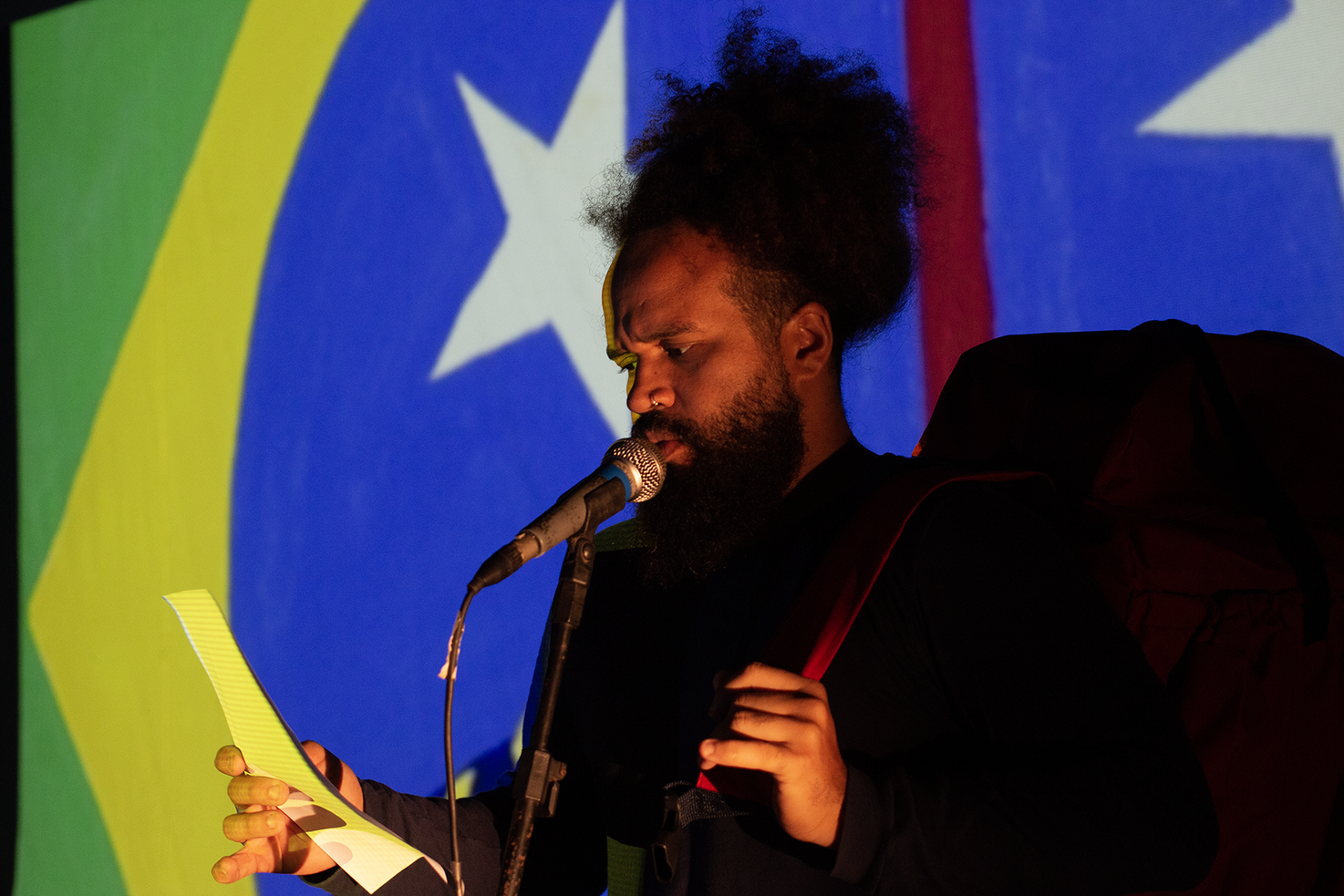 Photograph of Wellington Gadelha in front of a Brazilian flag he is speaking into a microphone.